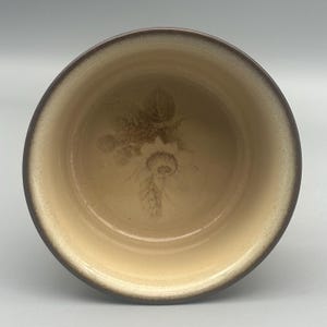 May include: A ceramic bowl with a floral design. The bowl has a light tan interior with a darker brown rim and exterior. A faded brown floral pattern is visible in the centre of the bowl.
