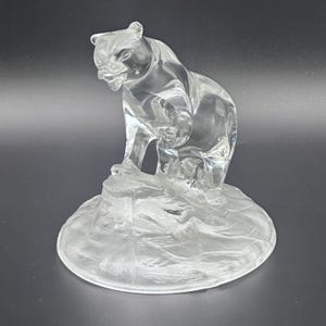 May include: A clear glass figurine depicting a bear standing on a textured base that resembles a rocky surface. The bear is captured in a dynamic pose with its head turned to the side, showcasing smooth, reflective surfaces and translucent details.