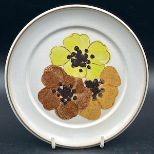 Può includere: Un piatto da portata in ceramica bianca con un bordo marrone e un motivo floreale con tre fiori in tonalità di giallo, marrone e arancione.