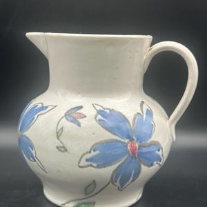 May include: Cream-colored ceramic pitcher with a rounded body and a handle. The pitcher is decorated with blue floral designs with red centers and gray outlines. The flowers are connected by green stems and leaves. The pitcher has a spout for pouring.