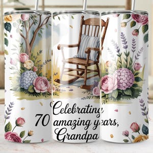 Op de afbeelding: Een witte beker met een bloemenontwerp en de tekst "Celebrating 70 amazing years, Grandpa." Het ontwerp omvat een houten schommelstoel, roze en paarse bloemen en een boom. De beker heeft een zilveren rietje.