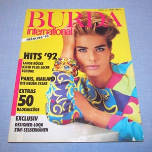 Op de afbeelding: Een cover van het Burda International magazine uit 1992. De cover toont een vrouw met blond haar en blauwe ogen. De achtergrond is geel, met de titel van het tijdschrift in roze en Duitse tekst. De vrouw draagt een kleurrijk jasje en een gouden armband.