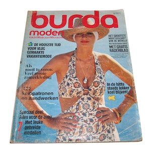 May include: A vintage Burda Moden magazine cover featuring a person wearing a straw hat and a patterned halter-neck swimsuit with a cutout design. The cover includes text in Dutch, such as 'burda moden', '122 knippatronen', and 'Met gratis raderblad'.