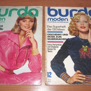 Op de afbeelding: Twee vintage Burda Moden tijdschriften. De covers tonen vrouwen in modieuze kleding. Het tijdschrift links heeft een roze outfit en rechts een blauwe top. De tijdschriften zijn uit de jaren 70.