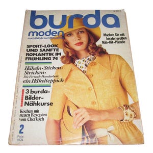 May include: Vintage February 1974 Burda Moden magazine cover. The cover features a woman in a yellow outfit and hat. Text includes "SPORT-LOOK UND SANFTE ROMANTIK IM FRÜHLING 74" and "3 burda-Bilder-Nähkurse".