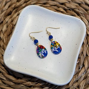 May include: A pair of dangle earrings with blue and gold accents. The earrings feature a teardrop-shaped charm with a colorful, hand-painted design.