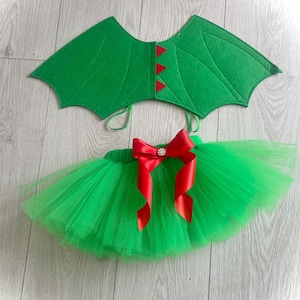 Puede incluir: Un conjunto de disfraz de dragón verde, que incluye una falda tutú con un lazo rojo y alas de fieltro verde. Las alas tienen detalles rojos y el lazo tiene una joya decorativa. Este disfraz infantil está diseñado para disfrazarse.