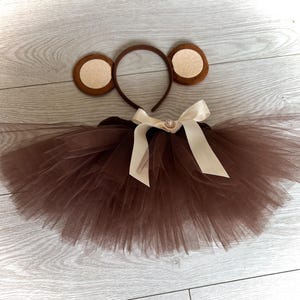 Può includere: Un set di costumi da scimmia marrone, composto da una gonna tutu, una fascia con orecchie e un fiocco. Il tutu è realizzato in tulle a strati e la fascia presenta orecchie marroni con centri beige. Il fiocco è color crema con un centro decorativo.