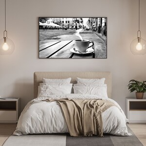 May include: A black and white photograph of a steaming cup of coffee on a wooden table in a city setting. The photo is hanging on a white wall above a bed with white bedding and a brown throw blanket.
