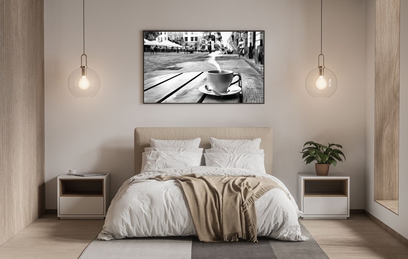 May include: A black and white photograph of a steaming cup of coffee on a wooden table in a city setting. The photo is hanging on a white wall above a bed with white bedding and a brown throw blanket.