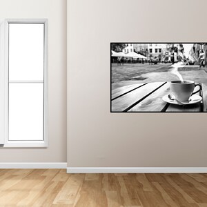 May include: Black and white photograph of a cup of coffee with steam rising from it, sitting on a wooden table in a city street. The background is blurred and shows buildings and people.