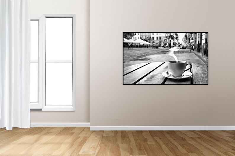 May include: Black and white photograph of a cup of coffee with steam rising from it, sitting on a wooden table in a city street. The background is blurred and shows buildings and people.