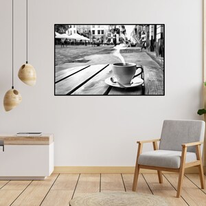 May include: A black and white photograph of a cup of coffee with steam rising from it, sitting on a wooden table outside a cafe. The cafe is bustling with people, and the photo is taken from a low angle, looking up at the cup.