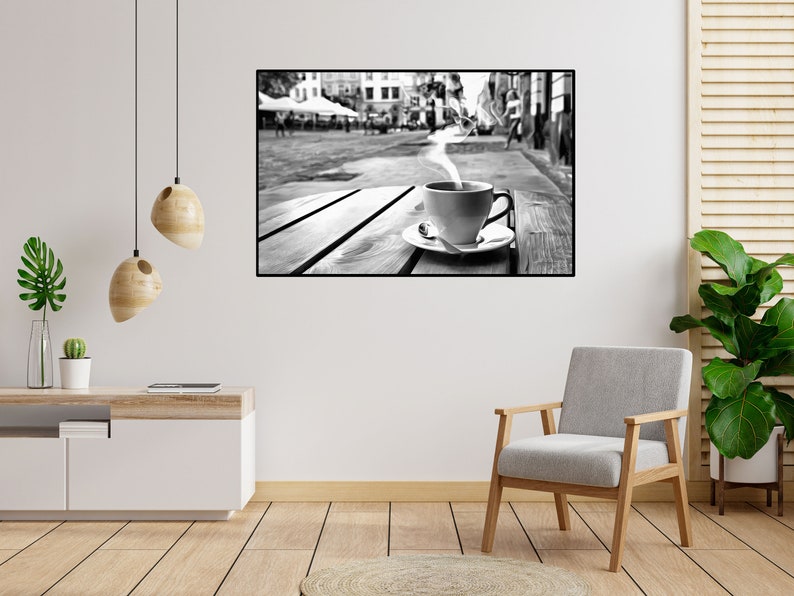 May include: A black and white photograph of a cup of coffee with steam rising from it, sitting on a wooden table outside a cafe. The cafe is bustling with people, and the photo is taken from a low angle, looking up at the cup.