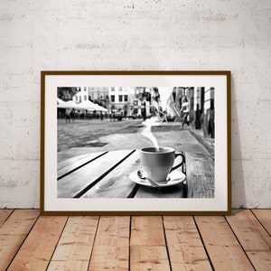 May include: A black and white photograph of a cup of coffee with steam rising from it, sitting on a wooden table outside a cafe. The cafe is in a city with buildings in the background.