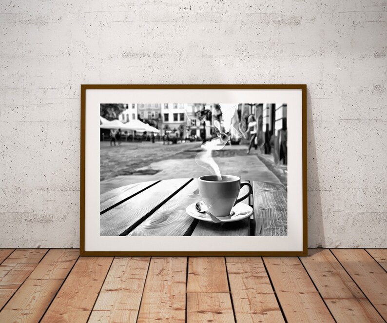May include: A black and white photograph of a cup of coffee with steam rising from it, sitting on a wooden table outside a cafe. The cafe is in a city with buildings in the background.