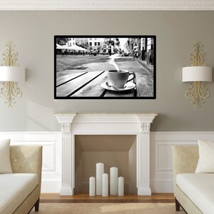 May include: Black and white photograph of a steaming cup of coffee on a wooden table in a city street. The cup is sitting on a white saucer. The photo is framed and hanging on a wall in a living room.