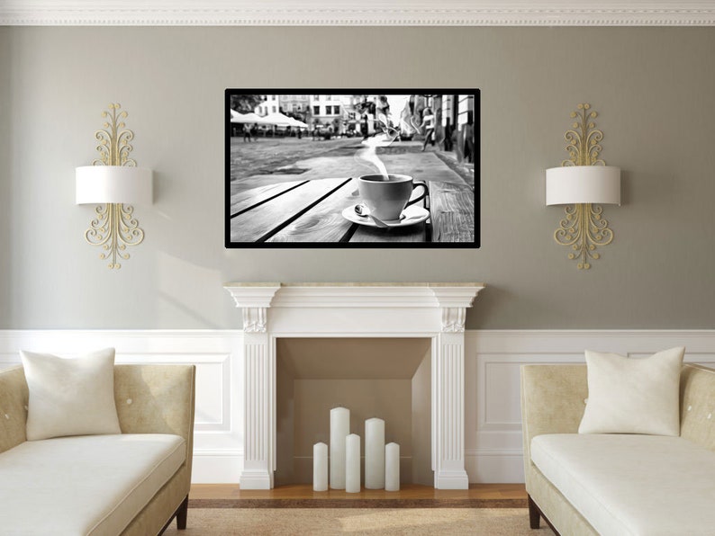May include: Black and white photograph of a steaming cup of coffee on a wooden table in a city street. The cup is sitting on a white saucer. The photo is framed and hanging on a wall in a living room.