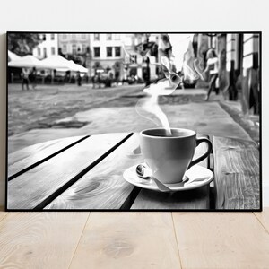 May include: A black and white photo of a steaming cup of coffee on a wooden table outside a cafe. The cafe is in a city with buildings and people in the background.
