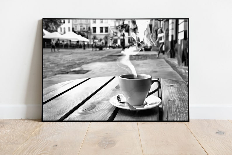 May include: A black and white photo of a steaming cup of coffee on a wooden table outside a cafe. The cafe is in a city with buildings and people in the background.