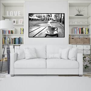 May include: A black and white photograph of a cup of coffee with steam rising from it, sitting on a wooden table in front of a city street scene.