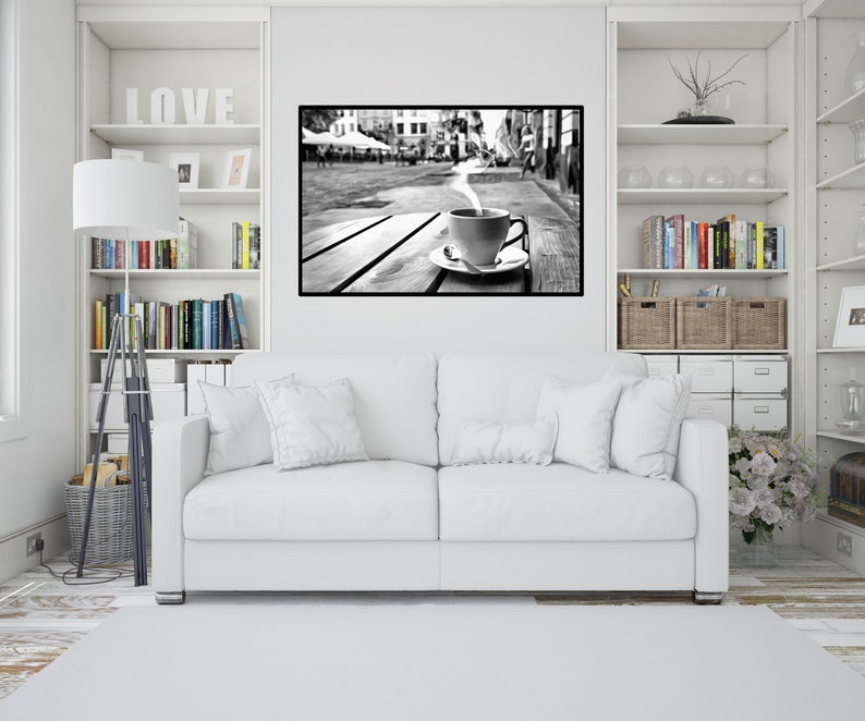 May include: A black and white photograph of a cup of coffee with steam rising from it, sitting on a wooden table in front of a city street scene.