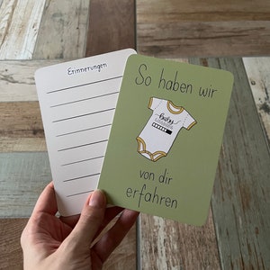 Può includere: Due carte, una bianca con la parola "Erinnerungen" e linee, l'altra verde con il testo "So haben wir von dir erfahren". La carta verde presenta un body per neonati con "baby loading".