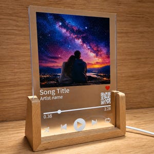 Könnte beinhalten: Personalisierte Musikplakette mit Holzsockel. Die transparente Acrylplatte zeigt ein romantisches Bild eines Paares unter einem Sternenhimmel. Die Plakette enthält Songtitel, Künstlername und eine Wiedergabeleiste mit QR-Code.