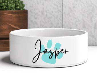 Könnte beinhalten: Weißer Keramik-Tiernapf mit einem türkisfarbenen Pfotenabdruck und dem Namen "Jasper" in schwarzer Schrift. Die Schüssel ist rund und steht auf einer weißen Oberfläche. Im Hintergrund befindet sich ein Holztopf mit einer grünen Pflanze.
