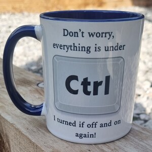 May include: White ceramic mug with a blue rim and handle. The mug has a humorous message that reads "Don't worry, everything is under Ctrl I turned it off and on again!"