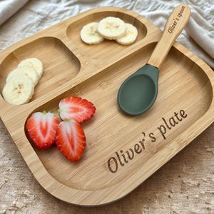 May include: A wooden divided plate with the text "Oliver's plate" and a matching spoon. The plate contains sliced bananas and strawberries. The spoon has a green silicone head and the text "Oliver's spoon".