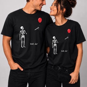 May include: A black crew neck t-shirt featuring a graphic of a white skeleton looking up at a detached skeletal hand holding a red balloon. The text "Well, shit..." is printed in white script below the skeleton on the front of the garment.