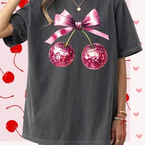 May include: An oversized charcoal grey t-shirt featuring a pink bow and two pink cherries with sequins. The cherries are on stems and the bow is tied around the stems.