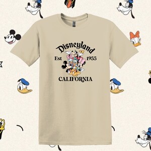 May include: A beige t-shirt with a black graphic of Mickey Mouse, Minnie Mouse, Donald Duck, Goofy, and Pluto. The graphic includes the text "Disneyland Est 1955 CALIFORNIA".
