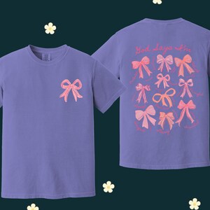 May include: Two purple t-shirts with pink bows printed on them. The front shirt has one bow and the back shirt has multiple bows with the text "Hot days are in" printed above them.