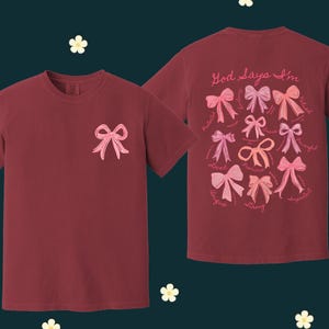 May include: Two maroon t-shirts with pink bows and the text "God Says I'm" on the front. The shirt on the right has a variety of pink bows with words like "loved", "strong", and "beautiful" around them.