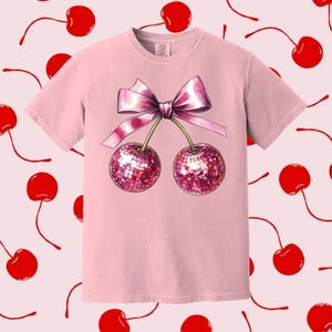 May include: Pink t-shirt featuring a graphic of two cherries with a pink bow. The cherries are sparkly and have a disco ball effect.