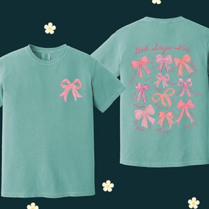 May include: Two teal blue t-shirts with pink bows embroidered on them. The front of one shirt has a single bow, while the back of the other shirt has multiple bows and the text "Good days are in".