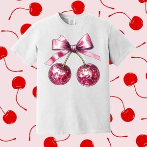 May include: White t-shirt featuring a pink bow and two cherries with sparkly red accents.