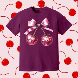 May include: A purple t-shirt featuring a pink bow and two cherries with sparkly red sequins. The cherries are connected by the bow.