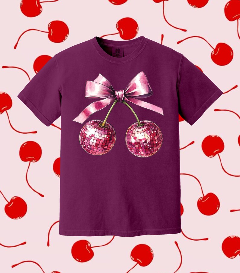 May include: A purple t-shirt featuring a pink bow and two cherries with sparkly red sequins. The cherries are connected by the bow.