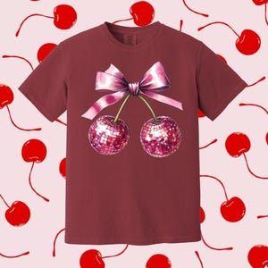 May include: A maroon t-shirt featuring a pink bow and two cherries with a sparkly design.