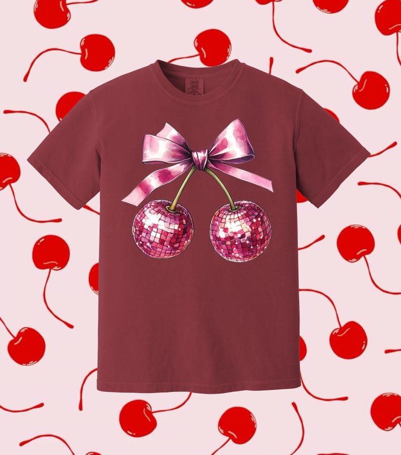 May include: A maroon t-shirt featuring a pink bow and two cherries with a sparkly design.