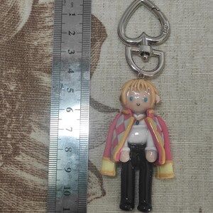 May include: A keychain featuring a miniature figurine of a character with blonde hair, blue eyes, and a pink and yellow jacket. The figure is approximately 2 inches tall, with a silver heart-shaped clasp.