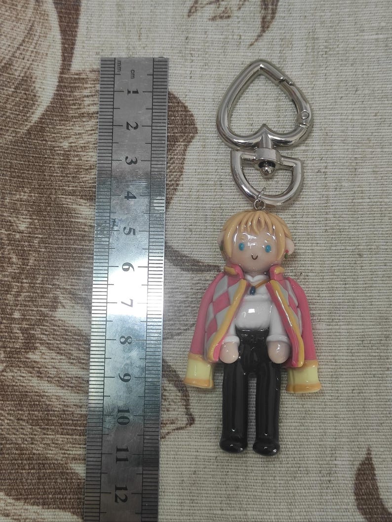 May include: A keychain featuring a miniature figurine of a character with blonde hair, blue eyes, and a pink and yellow jacket. The figure is approximately 2 inches tall, with a silver heart-shaped clasp.