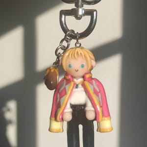 May include: A keychain featuring a miniature figurine of a character with blonde hair, blue eyes, and a pink and white jacket. The figure is wearing a white shirt, black pants, and black shoes. A small brown bag hangs from the keychain.