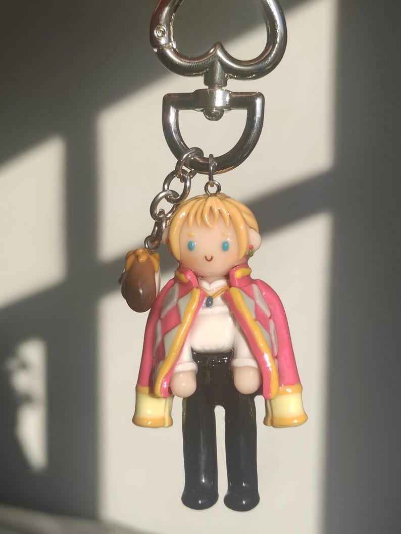 May include: A keychain featuring a miniature figurine of a character with blonde hair, blue eyes, and a pink and white jacket. The figure is wearing a white shirt, black pants, and black shoes. A small brown bag hangs from the keychain.