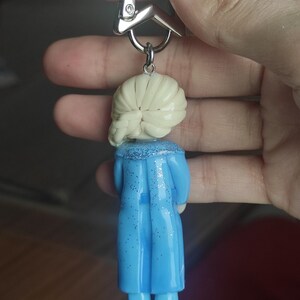 May include: A keychain featuring a miniature figurine of a character with blonde hair and a blue dress. The figurine is attached to a silver star-shaped clasp. The dress has glitter accents.