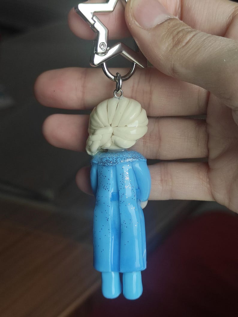 May include: A keychain featuring a miniature figurine of a character with blonde hair and a blue dress. The figurine is attached to a silver star-shaped clasp. The dress has glitter accents.