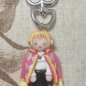 May include: A keychain featuring a cartoon figure with blonde hair, blue eyes, and a pink and white checkered jacket with yellow accents. The figure is wearing a white shirt and black pants. The keychain has a silver heart-shaped clasp.